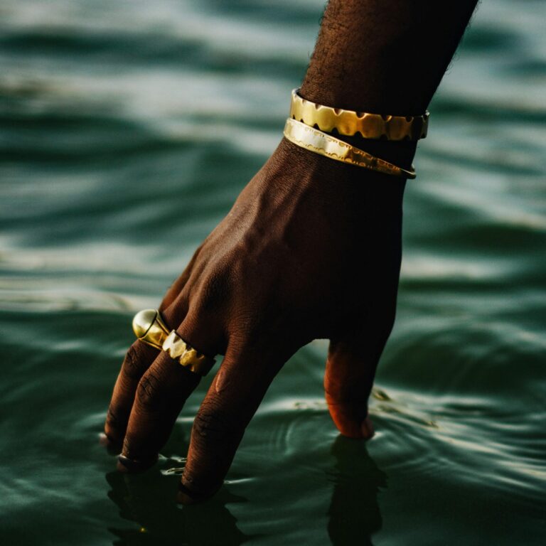 A hand adorned with gold jewelry gently touches calm water, creating a serene and luxurious atmosphere.