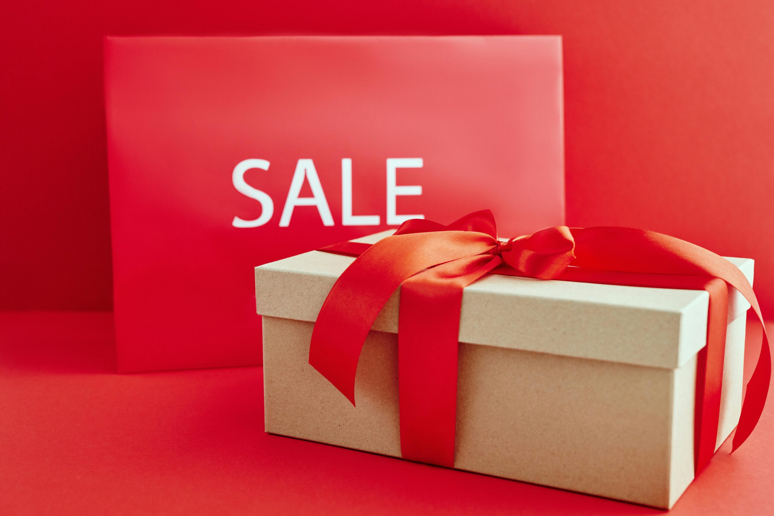 Gift box with red ribbon on red background with sale sign, perfect for retail promotions.