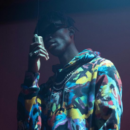 Black male musician in colorful hoodie holding a microphone performing on stage with dramatic lighting.